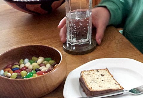 Esstisch, links eine Schale mit Geleebohnen, rechts ein Teller mit einem Stück Kuchen, oben eine Hand eines Teammitglieds, die ein Glas Mineralwasser hält und schräg dahinter eine Packung mit Salzstangen