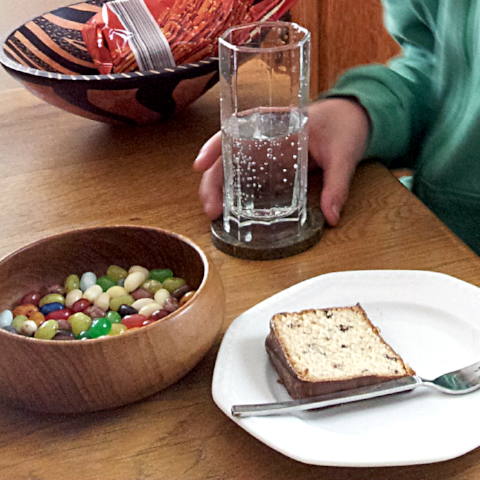 Esstisch, links eine Schale mit Geleebohnen, rechts ein Teller mit einem Stück Kuchen, oben eine Hand eines Teammitglieds, die ein Glas Mineralwasser hält und schräg dahinter eine Packung mit Salzstangen
